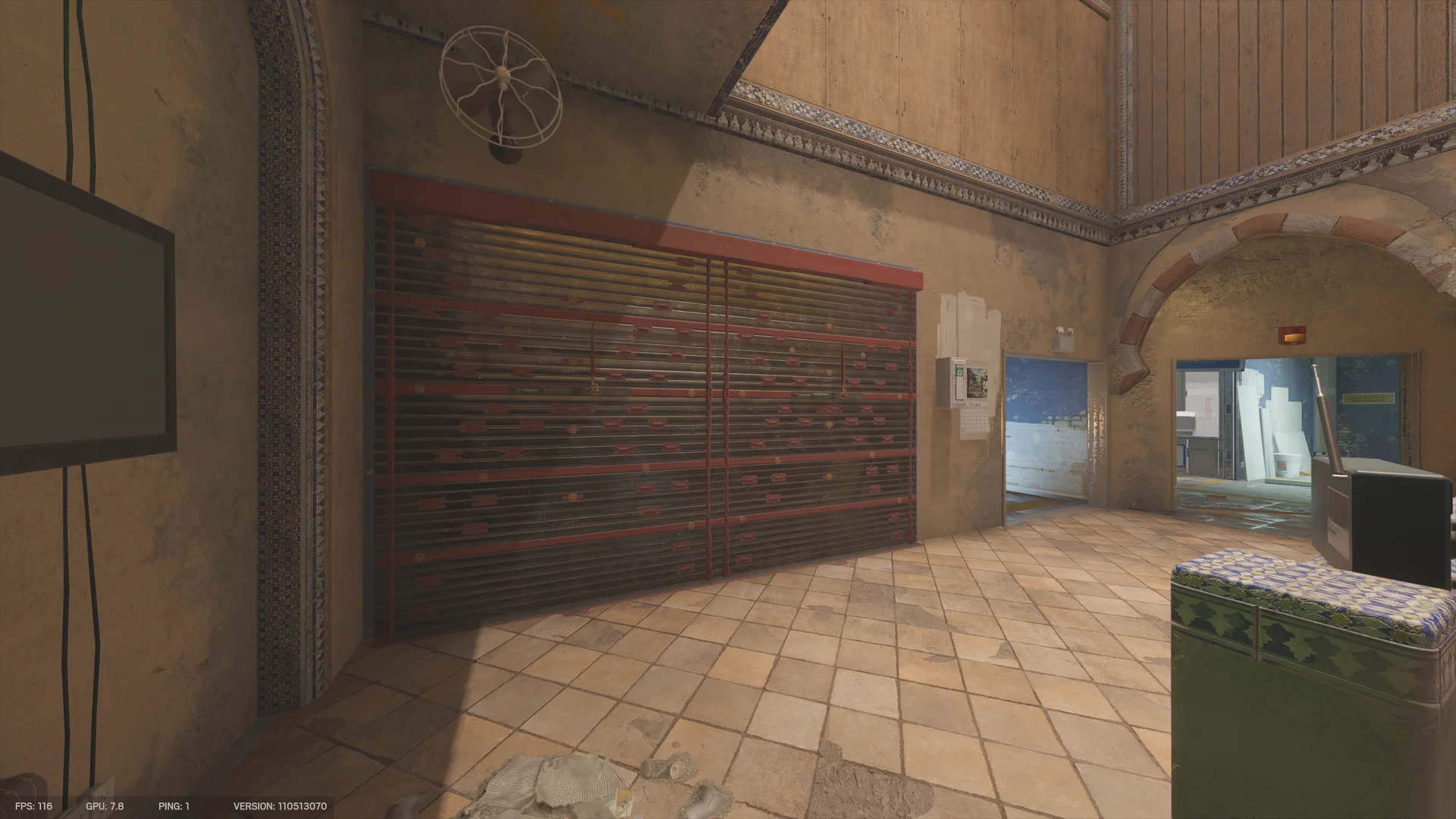 High-ceilinged tiled room on Border 1F with a wide closed metal shutter, a wall fan, a mounted TV at left, and doorways to blue-walled rooms ahead